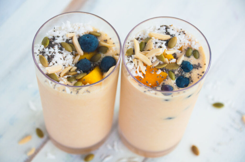 Two mango smoothies in tall glasses topped with blueberries, mango cubes, pumpkin seeds, almonds slices, coconut flakes, and chia seeds on a white background